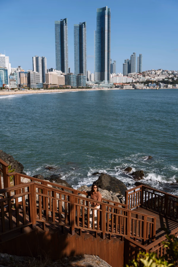 A woman standing on wooden steps in front of the sea with the glass towers of Busan in the background