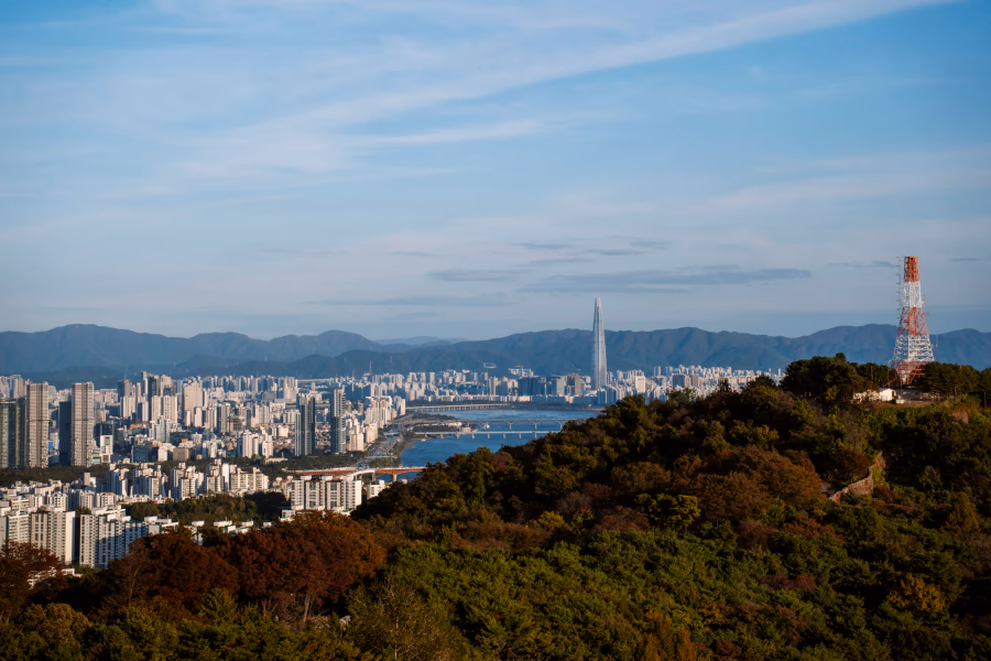 A large forested hill sits in front of the modern city skyline of Seoul South Korea backed by mounttains