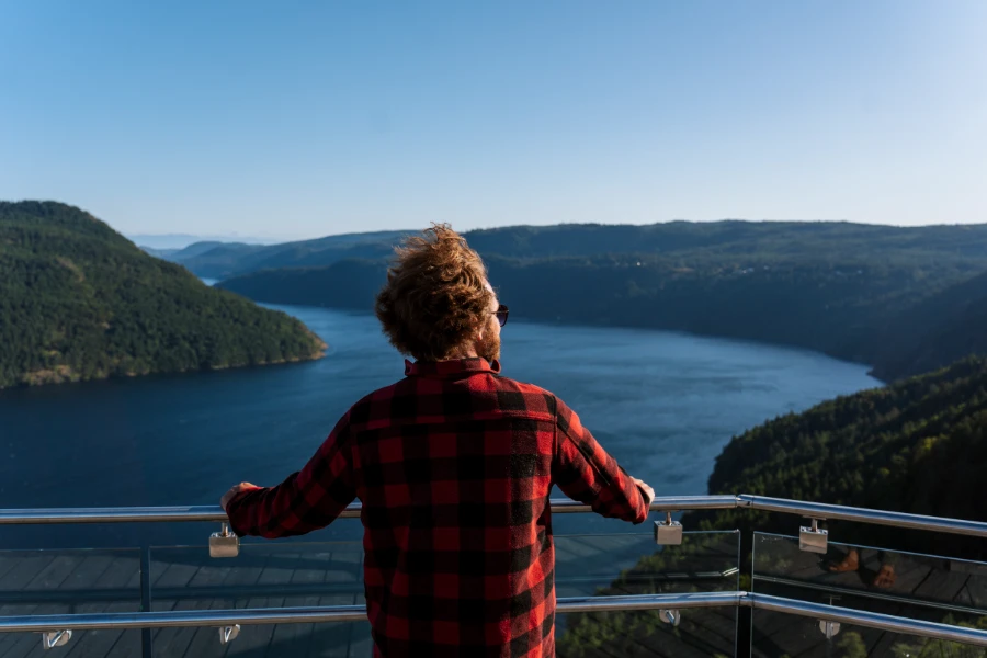 A man in a red flannel jacket stands with his hands on a glass and metal raining looking out to the Finlayson Arm from the top of the Malahat Skywalk