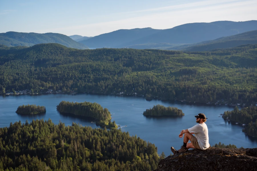 A man sits at the edge of a cliff at the top of Old Baldy Mountain above a deep blue lake with a vast green forest in the distance
