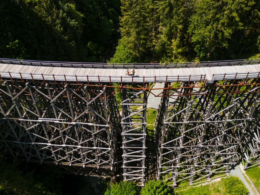 A man and woman walk hand in hand across a massive wooden trestle that spans across a lush canyon