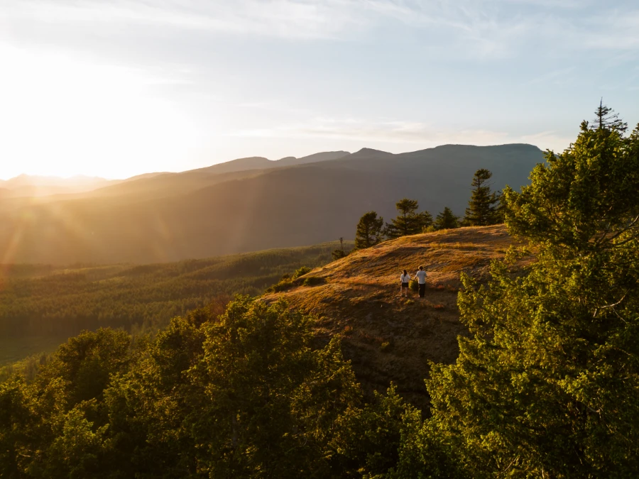 A man and a woman stand hand in hand at the top of Provost Mountain with a lush sea of green trees below them lit by the warm light of the setting sun