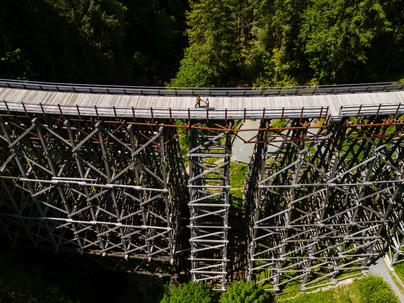 A man and woman walk hand in hand across a massive wooden trestle that spans across a lush canyon