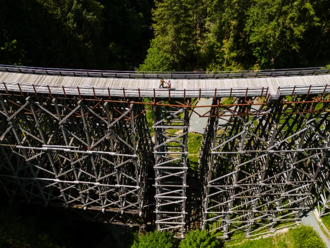 A man and woman walk hand in hand across a massive wooden trestle that spans across a lush canyon