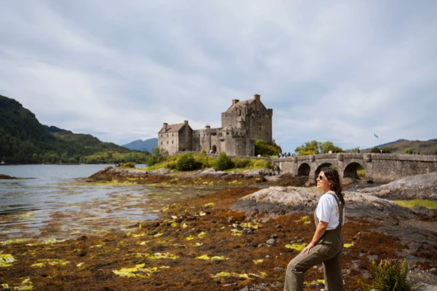 A woman standing in front of a long stone arched bridge that leads to a large stone castle