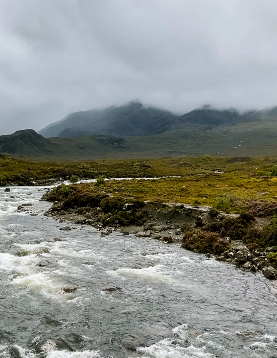 Water flows through a shallow river around a bend towards the mist shrouded mountains of the Isle of Skye
