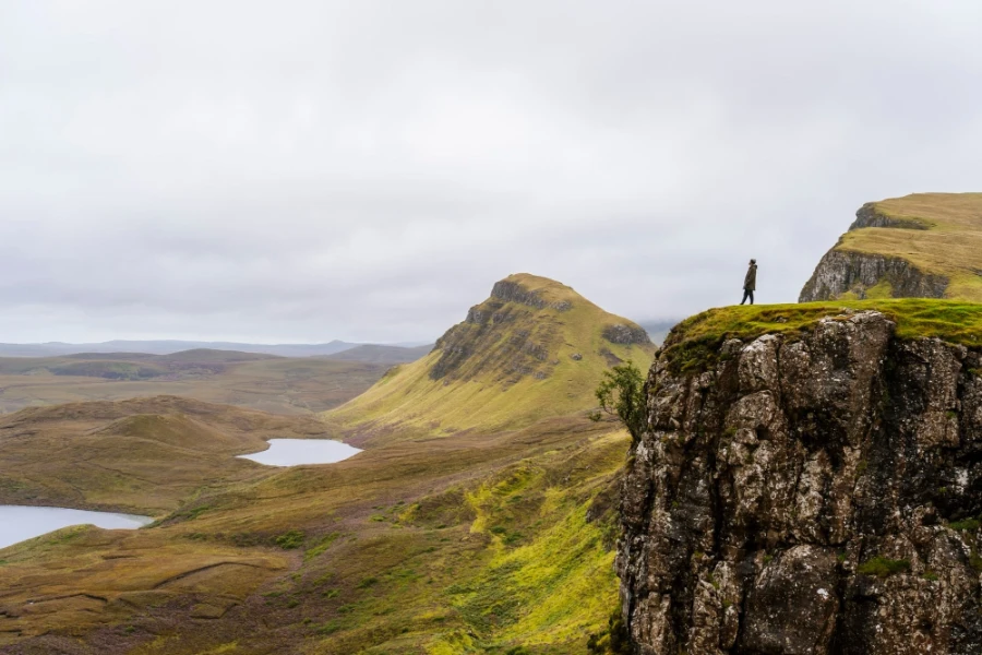 A man standing at the edge of a sheer cliff with a valley of rolling green hills stretched out beneath him