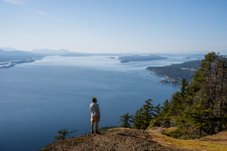 A man stands on the edge of a cliff high above the calm ocean with several Gulf Islands visible in the distance