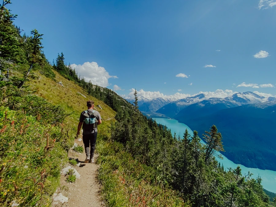 A man walks along a thin dirt path in Whistler Mountain alpine with a bright blue lake far below.