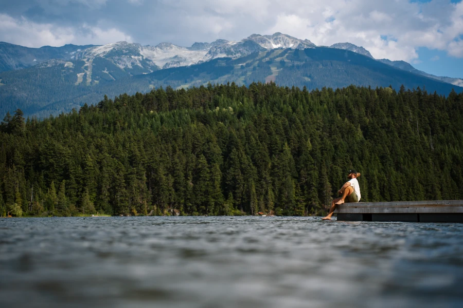 A man sits at the edge of a dock on Alta Lake in Whistler with rocky mountain peaks in the distance
