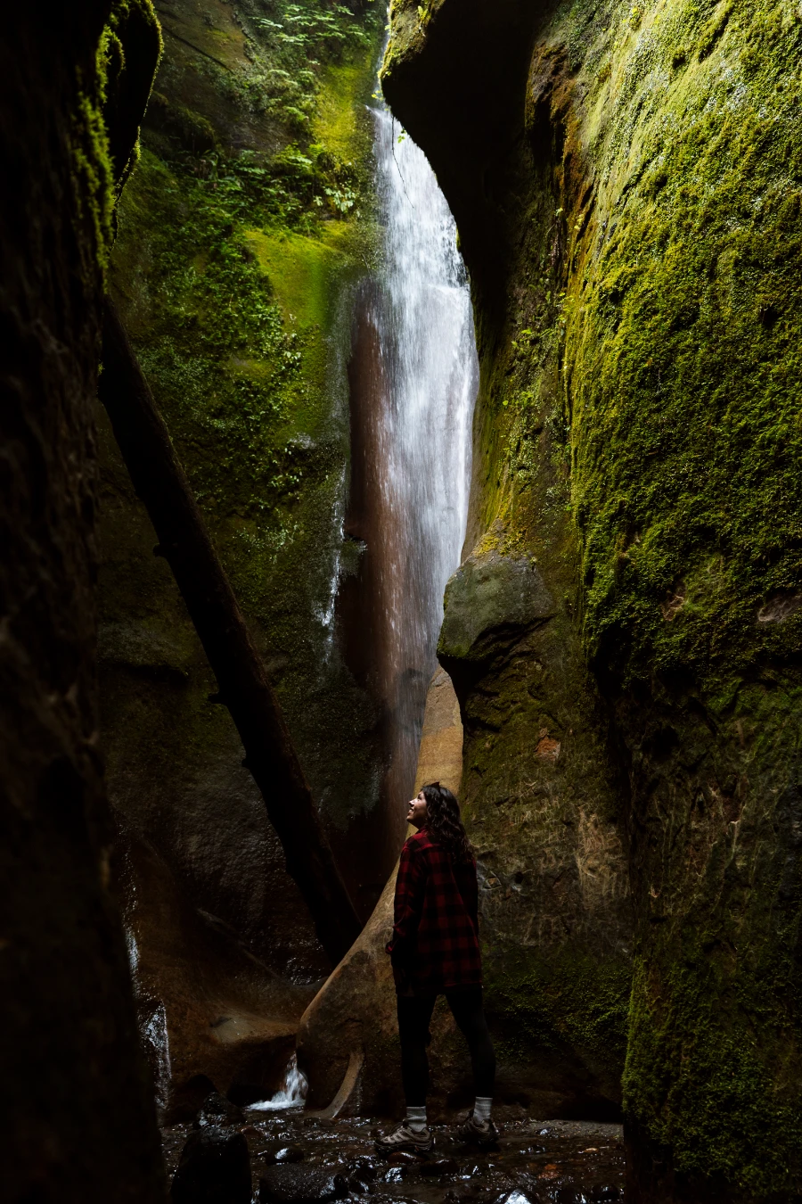 A woman walking through a dark, mossy cavern leads to a spot-lit waterfall at the end