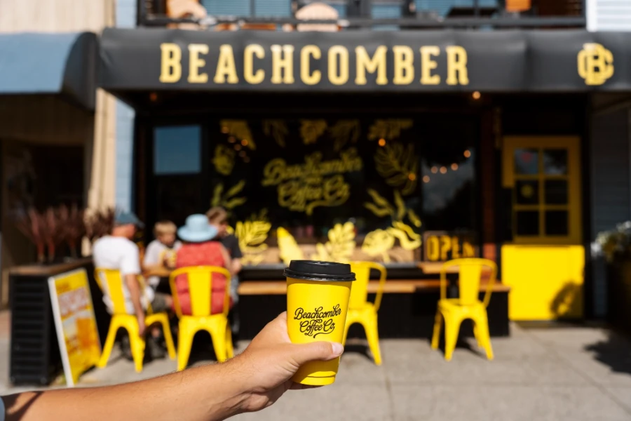 A hand hold a yellow coffee cup with a black lid up in front of a cafe with the same branded colors