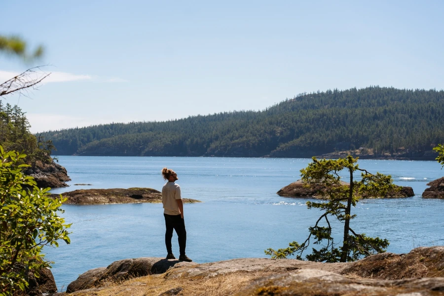 Man stands at the end of a large rock that is surrounded by calm sparkling water with a large tree covered island in the distance