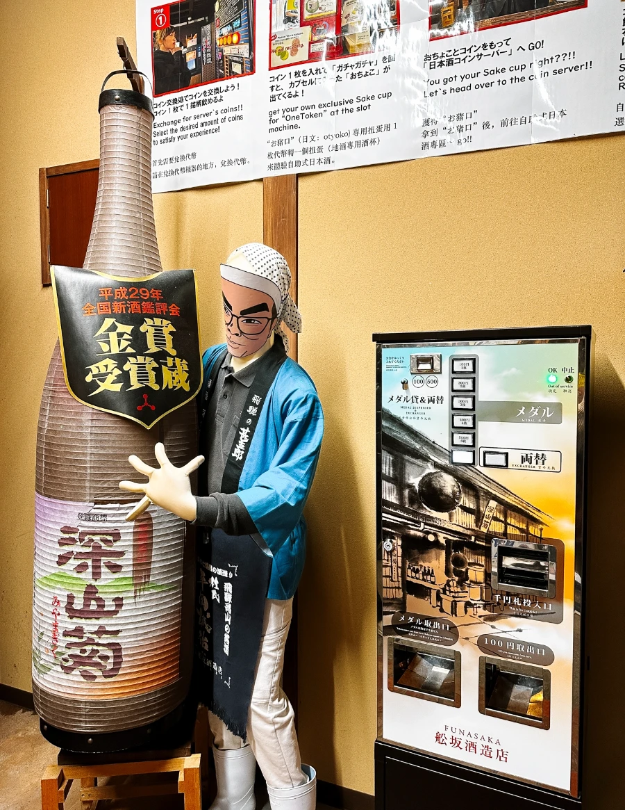 A mascot holds an oversized Sake bottle in front of a token dispenser machine in Takayama, Japan