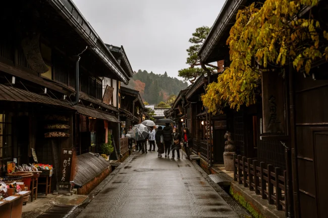 A crowd of tourists walk down a rainy street flanked by wooden storefronts 