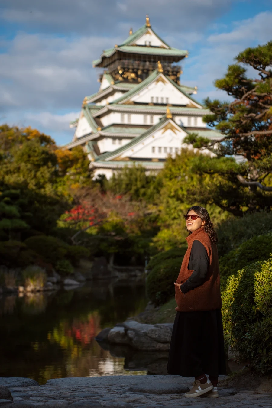 A woman stands in front of a calm pond with a multi story Japanese castle behind her