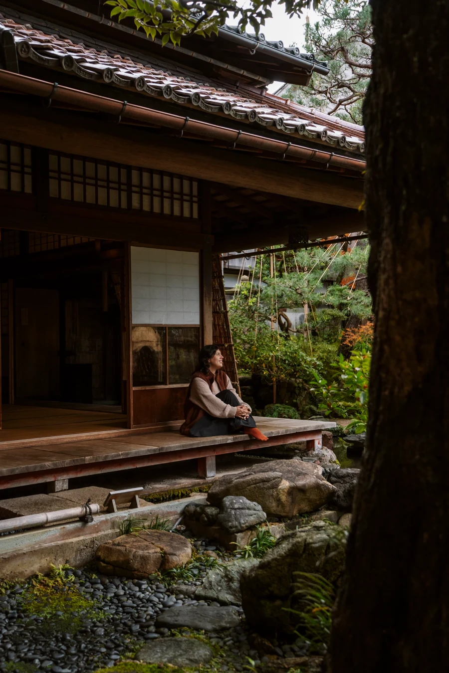 A woman sitting on a wooden deck looking out at a manicured garden with a traditional wooden Japanese house behind her