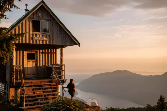 A woman stands next to a wooden hut and looks out to a wide view of forested valley with a large lake