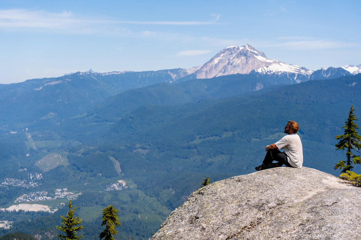 A man sitting at the edge of a stone cliff high above the Squamish Valley with Mount Garibaldi looming behind him.