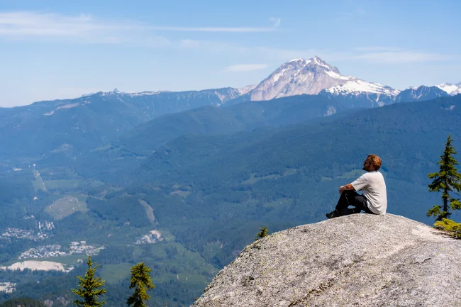A man sitting at the edge of a stone cliff high above the Squamish Valley with Mount Garibaldi looming behind him.