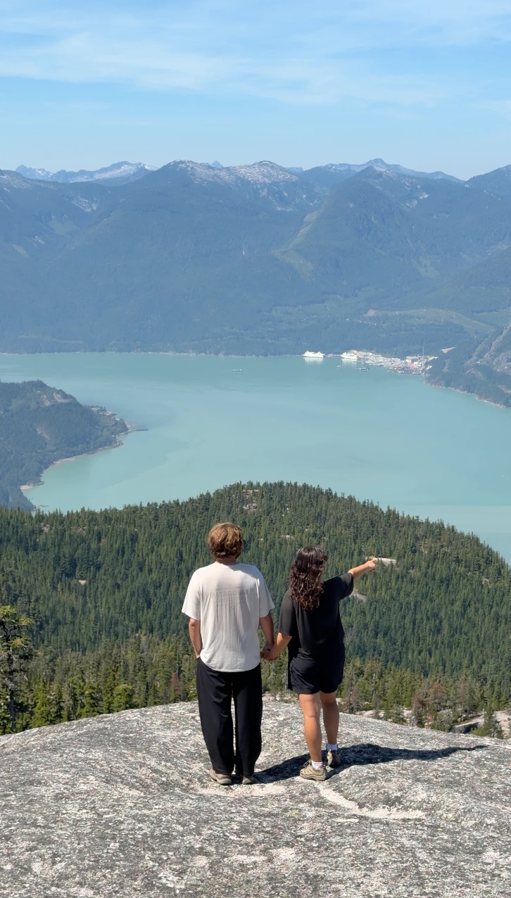 A man and a woman stand holding hands at the edge of a stone cliff with a large stretch of trees, the deep blue Howe Sound, and mountains beyond the water below them.