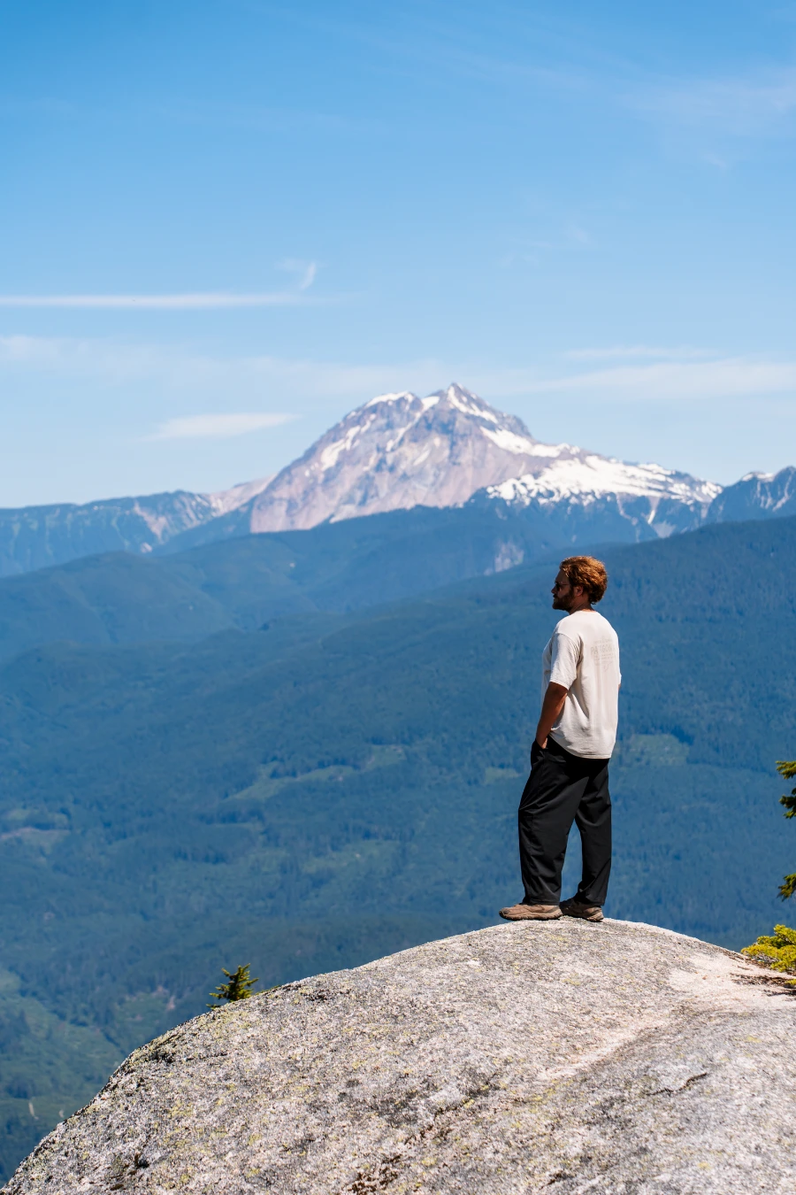 A man stands at the edge of a stone cliff high above the Squamish Valley with Mount Garibaldi looming behind him.