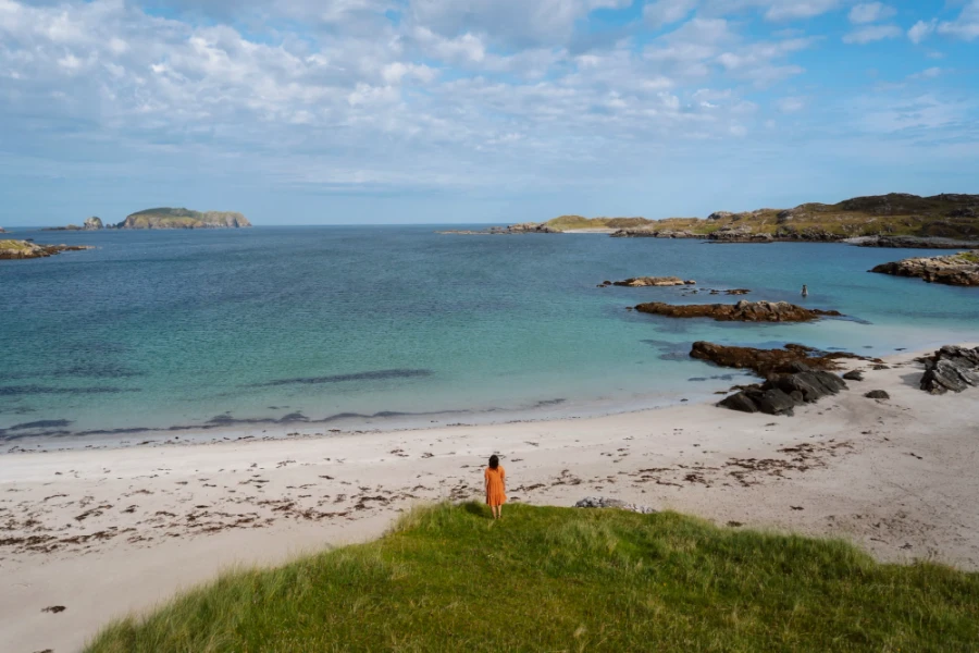 A woman in an orange dress stands at the end of a grassy patch looking down at the white sand of Bosta Beach, Isle of Lewis