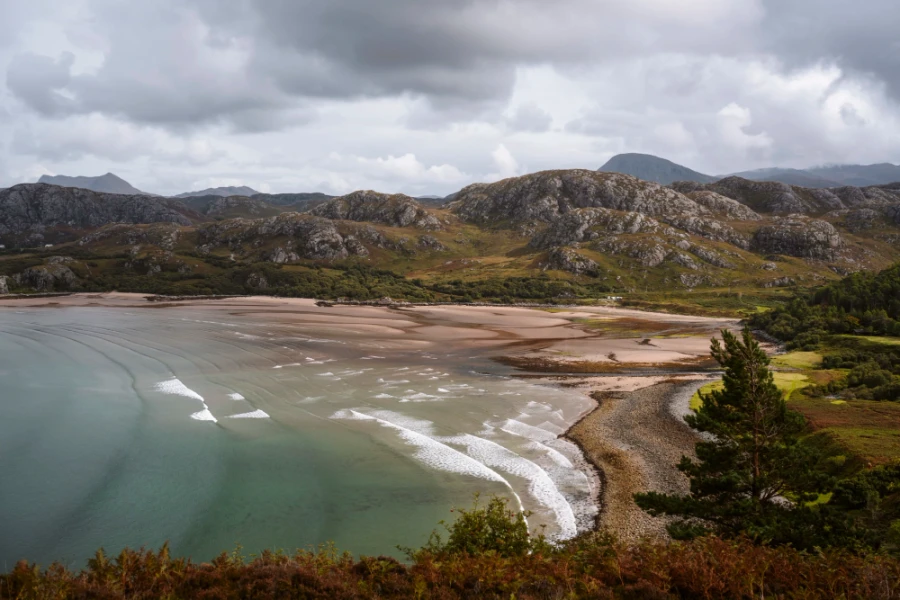 Large white wash waves break along the sand shores that sit below rolling stone hills at Gruinard Beach, Torridon