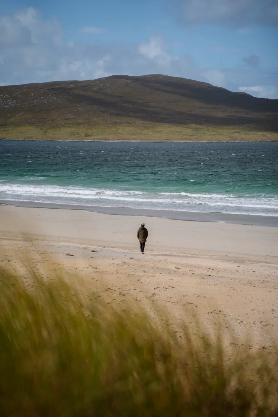A man in a large jacket is walking  out onto the beach towards a calm sea on Seilebost Beach, Isle of Harris