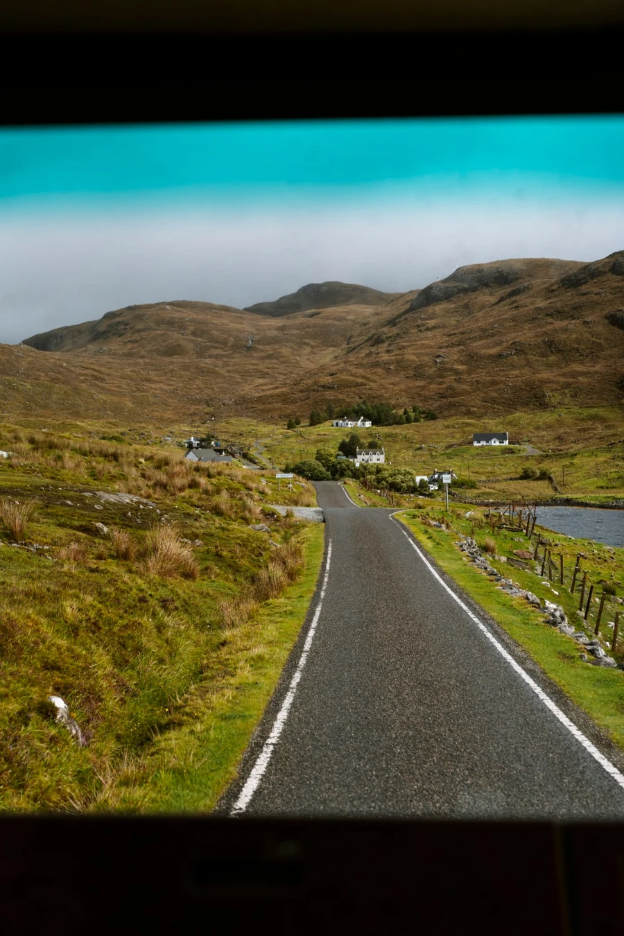 A single track road in Scotland that leads to several cottages and is framed through the windshield of a vehicle