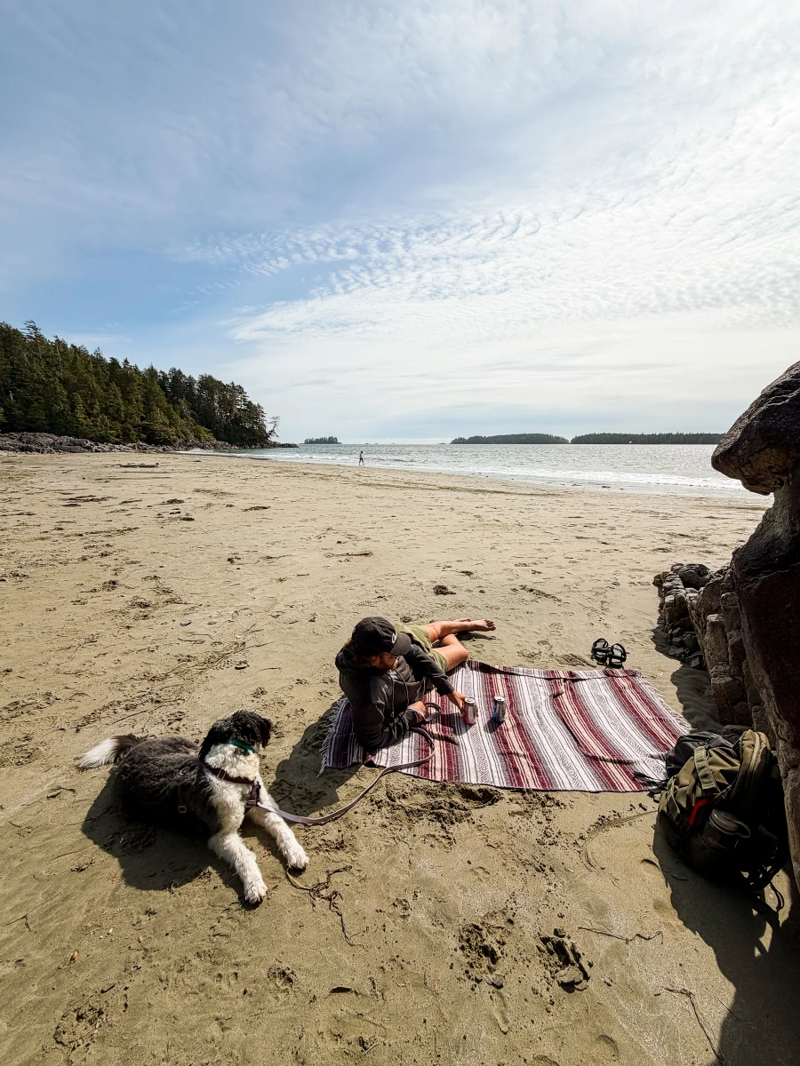 A man lays on a picnic blanket on a sandy beach with his dog at low tide at Tonquin Beach in Tofino