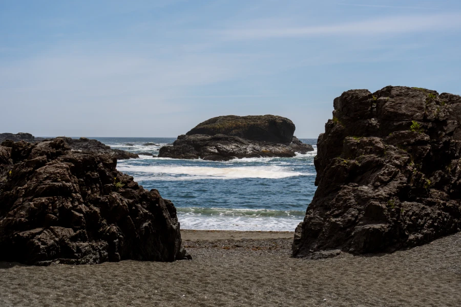 Two large rocks on a sandy beach frame a third large rock that is further out in the water as frothy white waves crash into the framed rock and the shore past the framing rocks