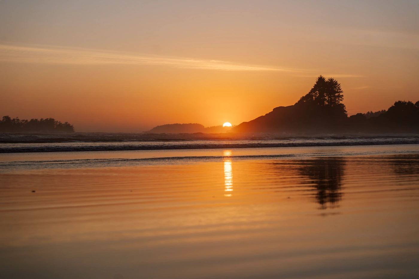 The wet sand of tin wis beach at low tide glows golden as it reflects the warm light of the setting sun the is dipping below the tree-lined horizon