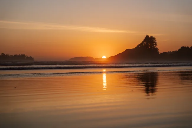 The wet sand of tin wis beach at low tide glows golden as it reflects the warm light of the setting sun the is dipping below the tree-lined horizon