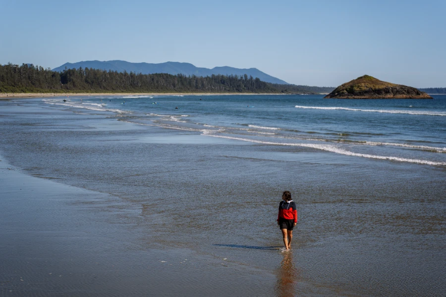 A woman wearing shorts and a wind-breaker jacket walks in ankle deep water along the sandy shores of Long Beach near Tofino