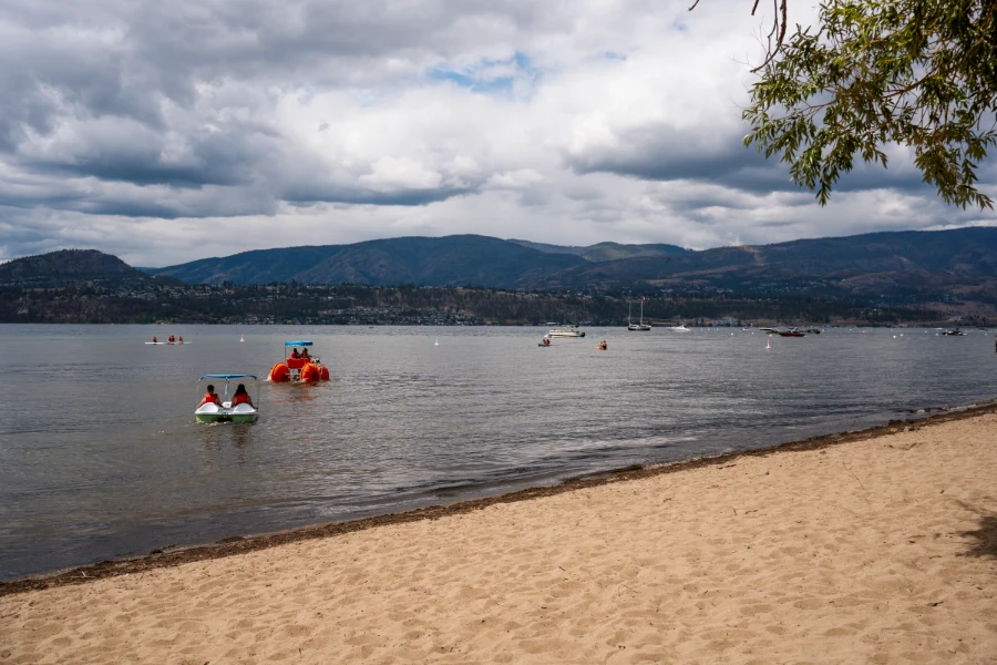 A white sandy beach on the edge of Okanagan Lake with a paddle boat and an odd watercraft with massive wheels floating just offshore