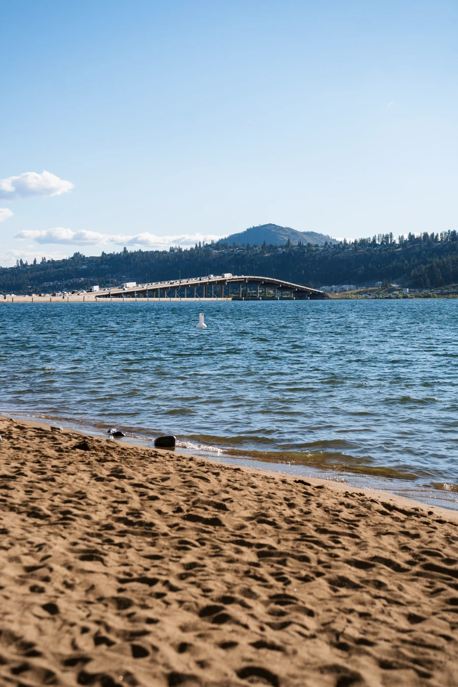 A small strip of sandy beach on the shore of Okanagan Lake with the iconic William R. Bennett Bridge visible in the distance