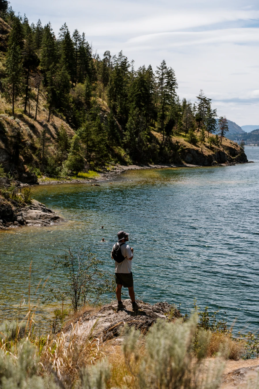 A man stands at the edge of a cliff above the swimming area at Paul’s Tomb with a tree-covered shoreline stretching past him into the distance