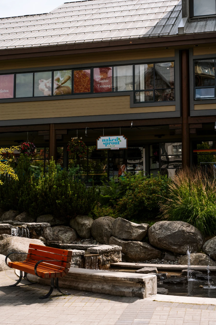 A sign for the Naked Spout hanging along a shaded walkway behind a public fountain in Whistler Village