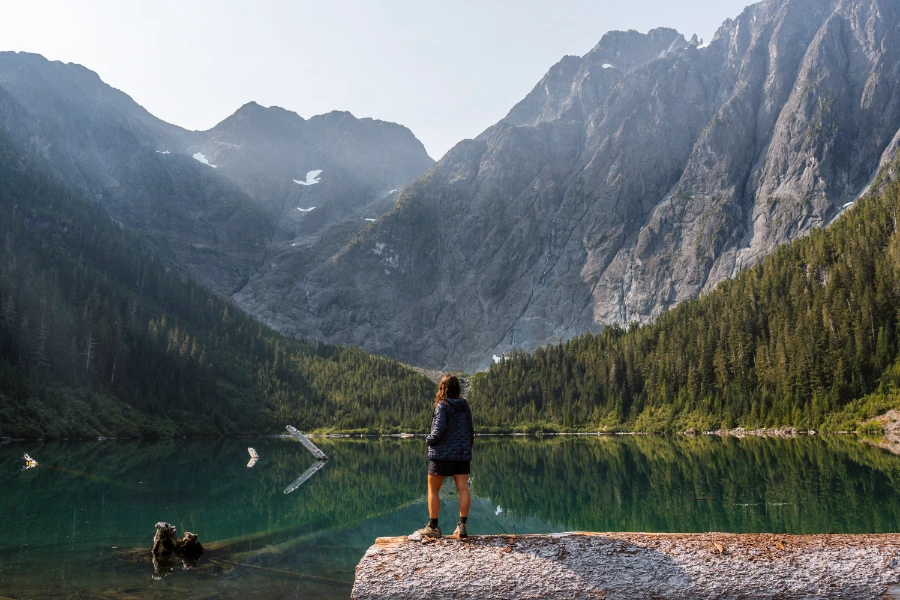A woman stands on a massive log next to a lake with a clear reflection of the massive jagged mountains in the distance