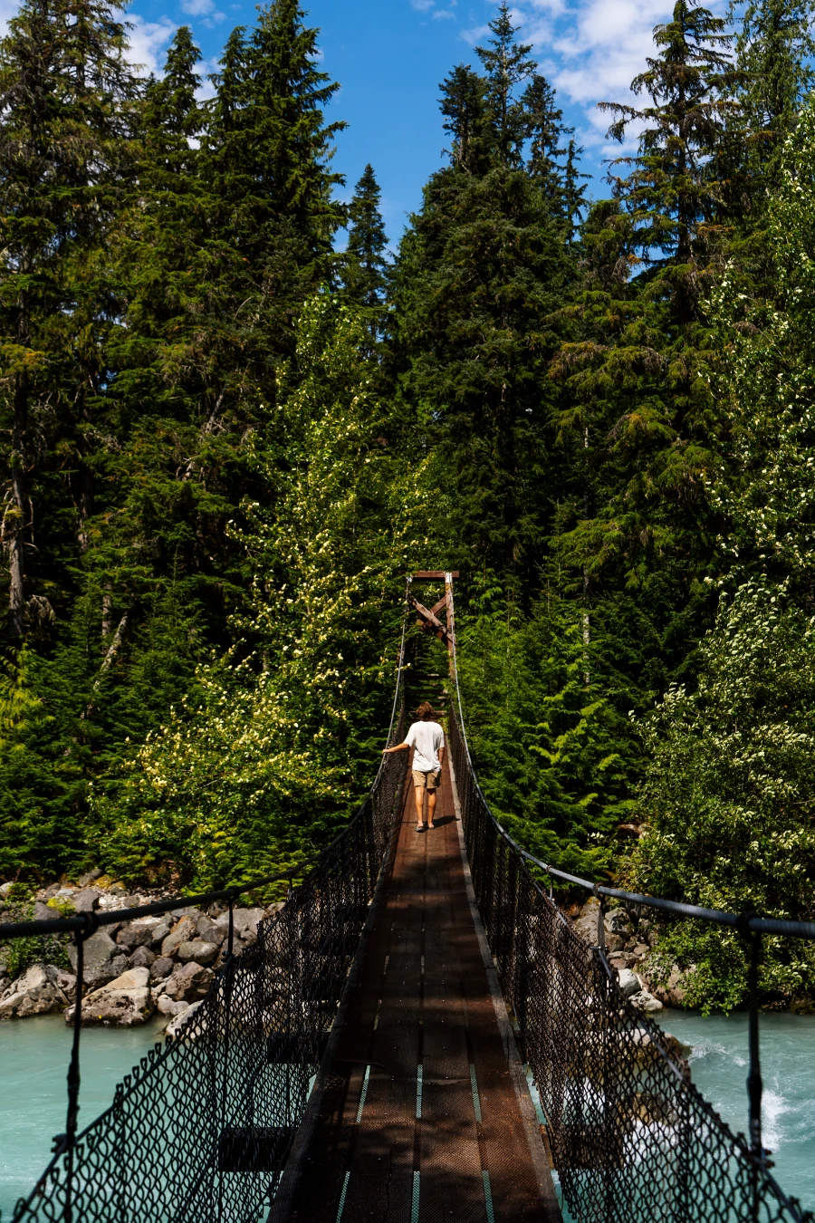 A man walks across a narrow suspension bridge that crosses the fast-moving and strikingly blue water of Cheakamus River
