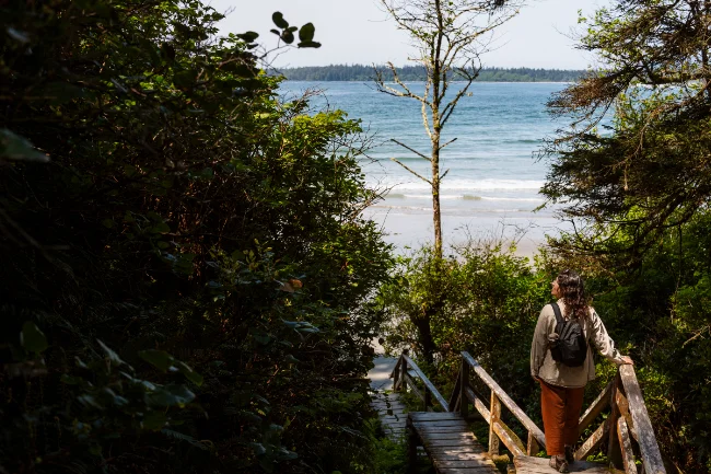A woman walking down a large boardwalk staircase passing by lush shrubery leading down to a wide sandy beach in Tofino, BC