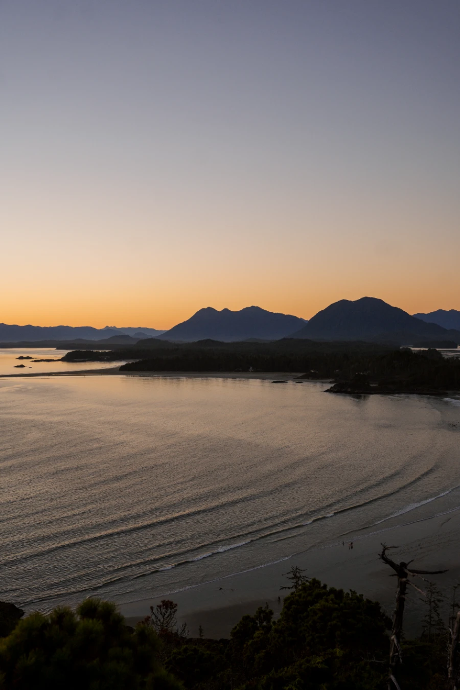 Gentle rows of waves roll into the tree-lined cove of Cox Bay with mountains on the horizon in silhouette at sunset