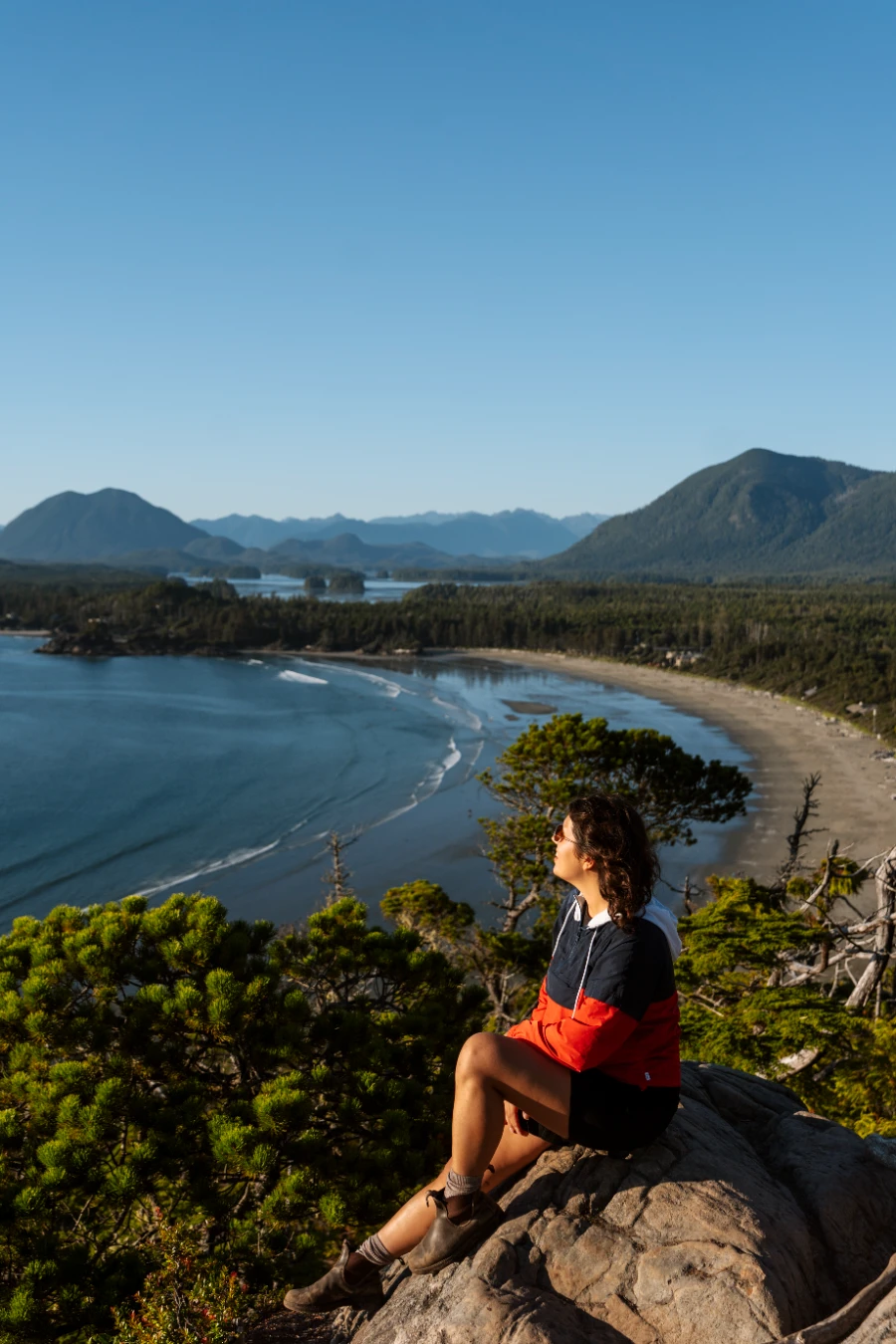 A woman sitting on a rock high above Cox Bay in Tofino with gentle waves rolling into the sandy shore far below her