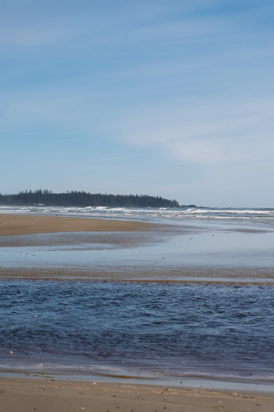 A large tidal river flows across the sand of Combers beach into the sea where rough waves batter the sandy shore that stretches far into the distance