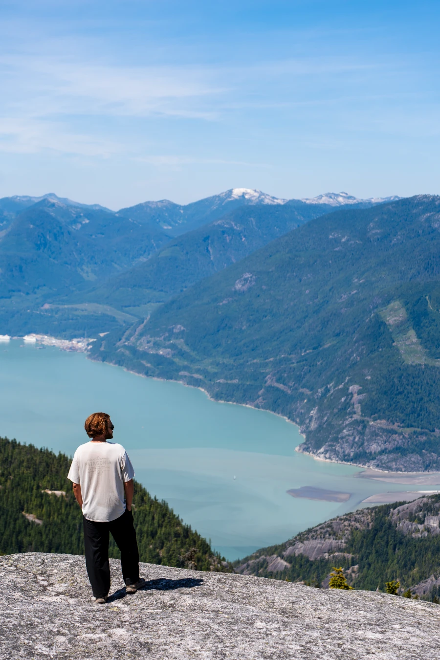 A man stands at the edge of a bare stone cliff high above the deep blue waters of the Howe Sound.