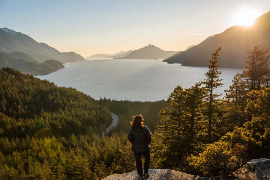 A woman stands at the end of a rocky cliff, standing above a sea of green trees that are growing between the Sea-to-Sky Highway winding through the valley on one side, and the deep blue waters of the Howe Sound on the other.