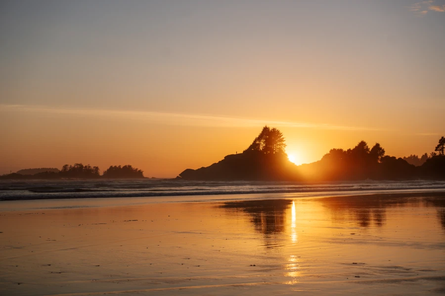 The wet sand reflects the warm sunset light at Cox Bay Beach while the sun sets behind a distant rocky outcrop