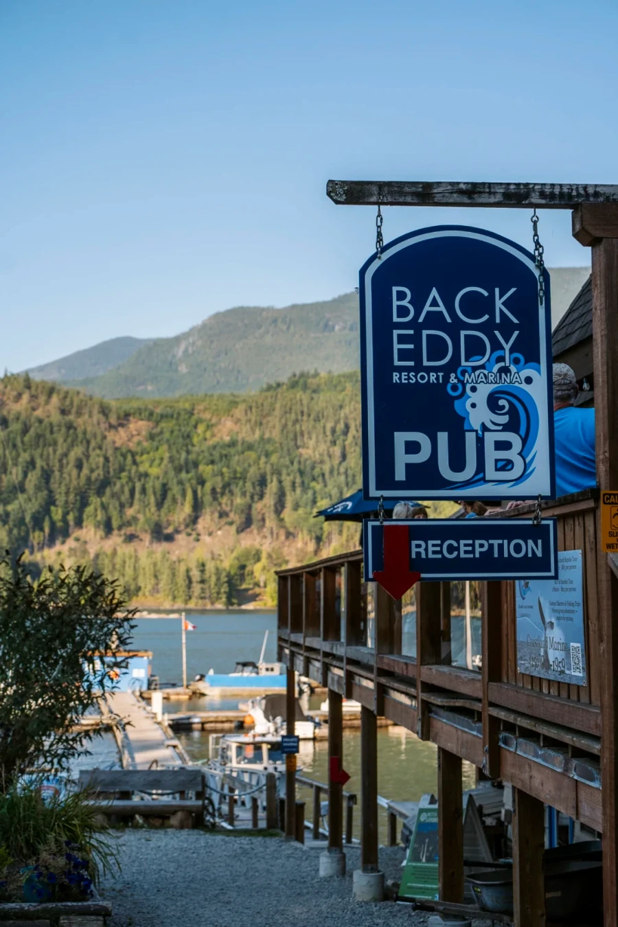 Welcoming sign for the Back Eddy Pub reception hangs with docked boats on the water in the distance