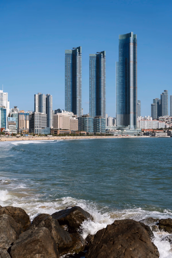 Waves crash onto large rocks with the long stretch of white sand at Haeundae Beach in the distance being bordered by several large glass towers